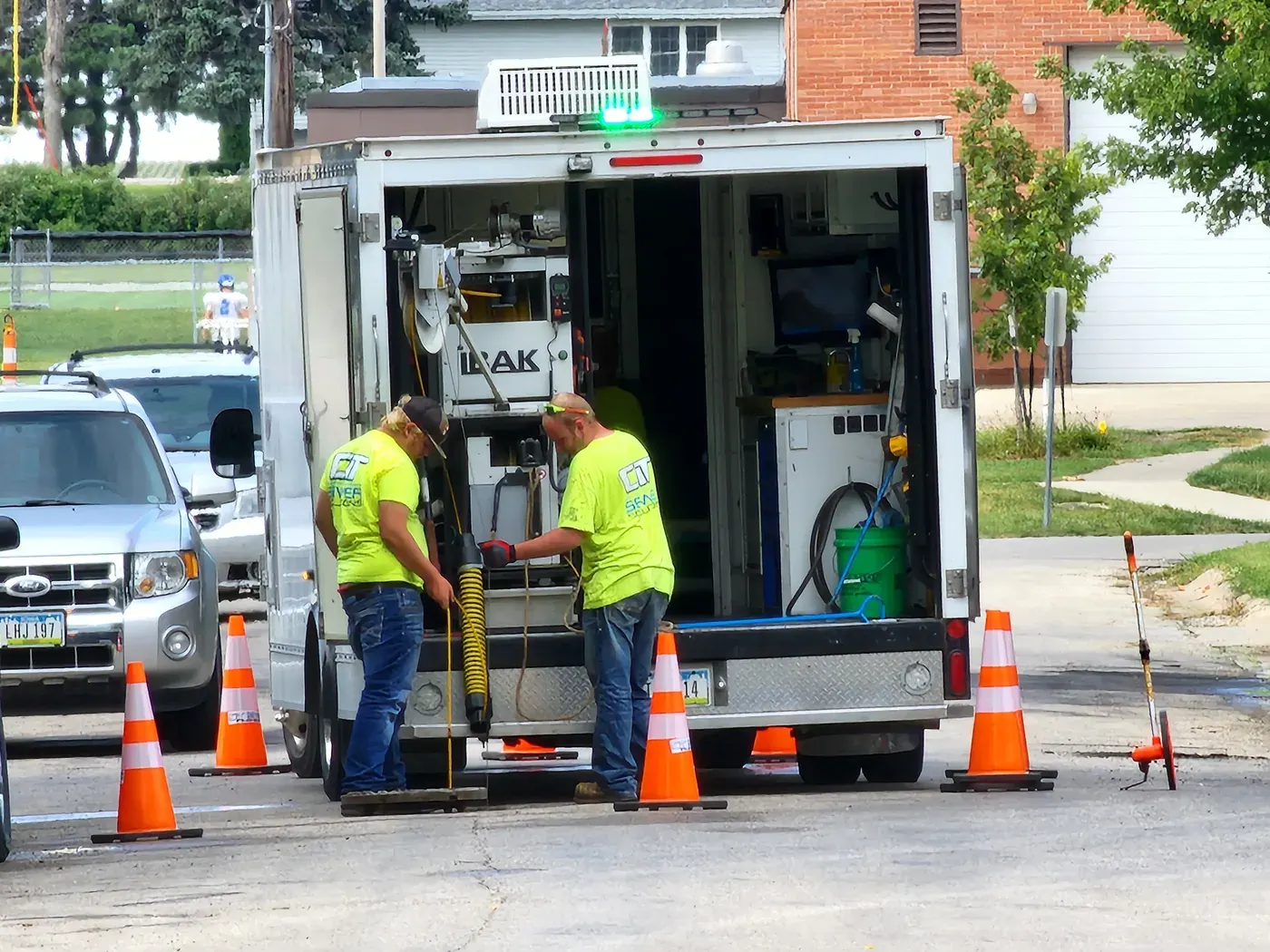 Two CIT crew members stand beside an open manhole at the rear of an inspection truck, with cable equipment and traffic cones visible.